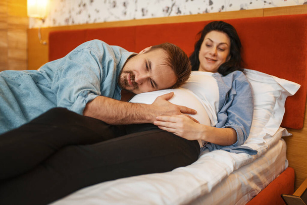 Husband listens to baby in pregnant wife's stomach at home, bedroom interior on background. Pregnancy, prenatal period. Expectant mom and dad are resting on sofa, health care - Photo, Image