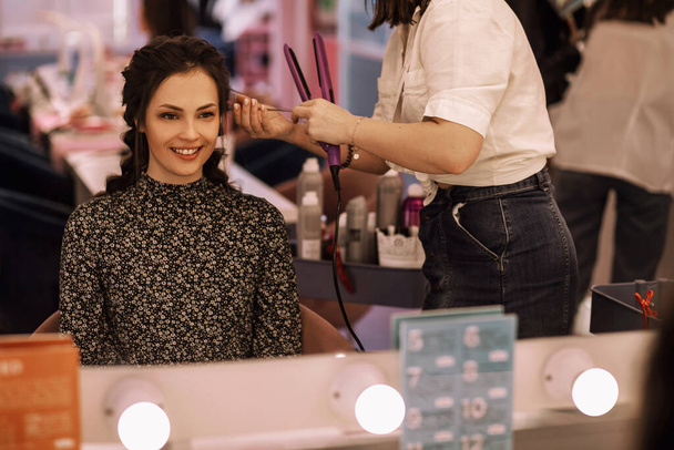 young woman in the women's salon uses the services of a hairdresser, a professional hairdresser straightens her hair with a curling iron, the girl smiles and is satisfied with the work of the stylist - Photo, Image