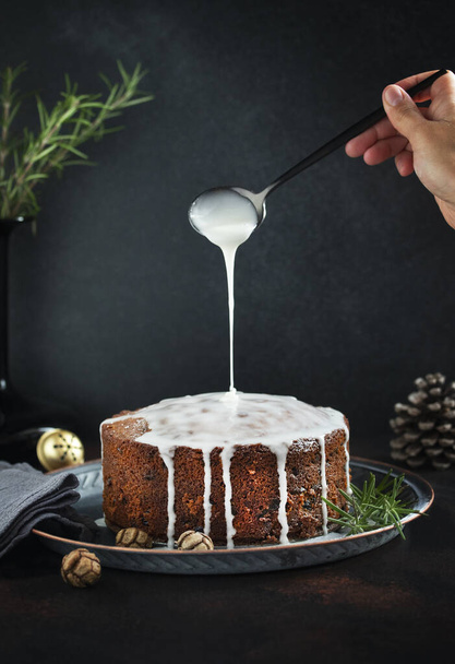 Традиционный рождественский торт с фруктами и орехами на темном столе и черном фоне. Женская рука поливает торт сахаром из глазури. Десерт на зимние каникулы. По вертикали. Копирование пространства - Фото, изображение
