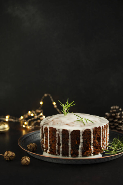 Traditional Christmas cake with fruits and nuts on a dark table and black background. Dessert for the winter holidays. Christmas decorations and lights. Copy space - Photo, Image
