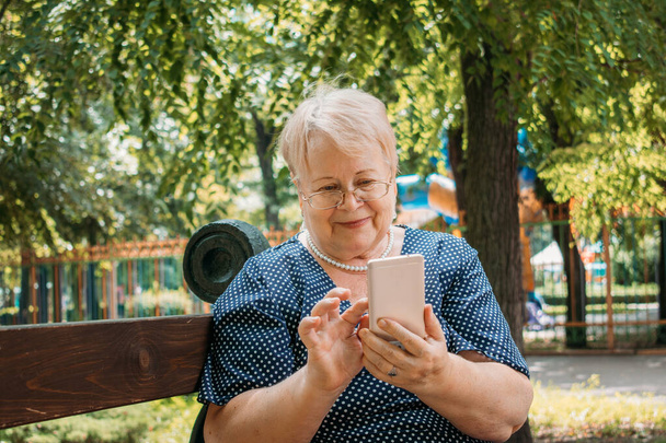 公園でスマートフォンを持つ陽気な老婦人。携帯電話を使用して公園で幸せなシニア女性の屋外肖像画。プラスサイズブロンド高齢女性持っていますビデオ通話. - 写真・画像