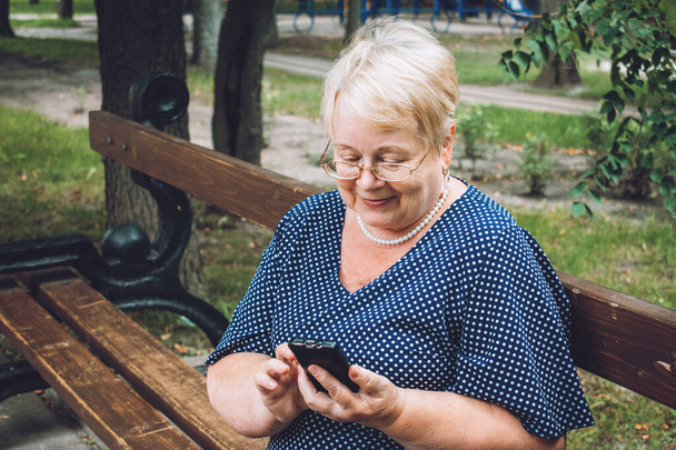 Veselá stařena s chytrým telefonem v parku. Venkovní portrét šťastné starší ženy v parku pomocí mobilního telefonu. Plus velikost blond starší žena mají video hovor. - Fotografie, Obrázek