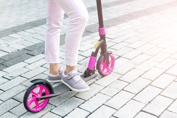 Bottom part of young adult female person wearing pink casual jeans and sneakers riding push kick scooter at city street on summer day outdoor. Teenager girl having fun enjoy sport activities outside. - Photo, Image