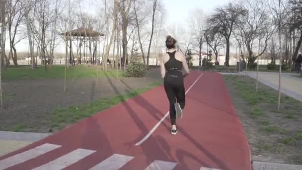 Młoda kobieta biegnie po torze w parku. Aktywność, człowieku. Biegacz biegnie po torze. Utrzymywanie ciała w doskonałym stanie. Tor z gumową opaską do chodzenia i biegania wyścigowego - Materiał filmowy, wideo