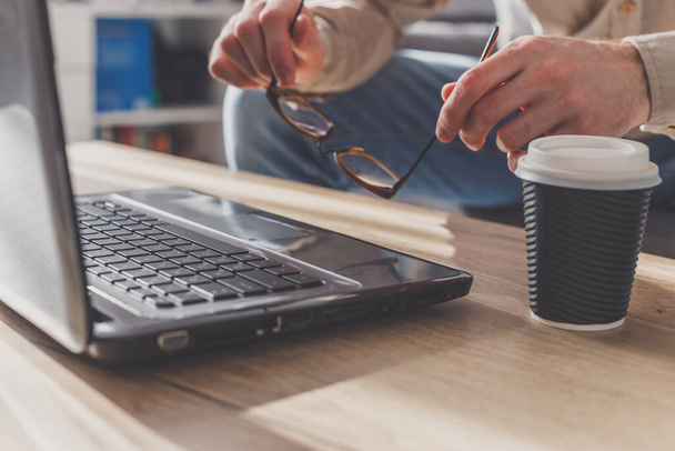 Mann setzt Brille auf Laptop, um eine kurze Pause zu machen, müde nach langer Arbeit oder nervös. Kaffeetasse auf Tisch neben Laptop. - Foto, Bild