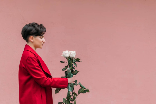 A nemek közötti egyenlőség, a nők napja koncepció. Szabadtéri portré fiatal barna nő rövid frizura piros kabátban gazdaság csokor fehér rózsa rózsaszín fal háttér. - Fotó, kép