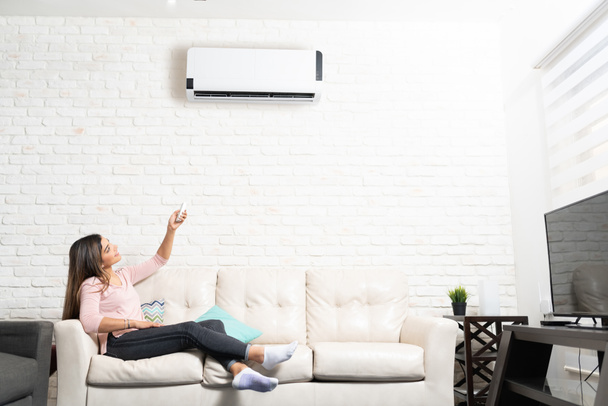 Latina mulher ligar ar condicionado unidade usando controle remoto enquanto sentado no sofá em casa
 - Foto, Imagem