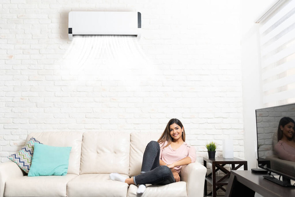 Smiling Hispanic beautiful woman resting on sofa below air conditioner at home - Fotoğraf, Görsel