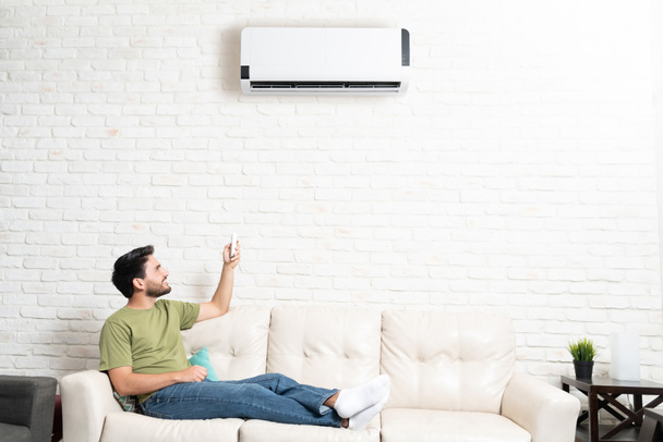 Volledige lengte van de Latijnse man het inschakelen van airconditioner met behulp van afstandsbediening tijdens het zitten op de bank thuis - Foto, afbeelding