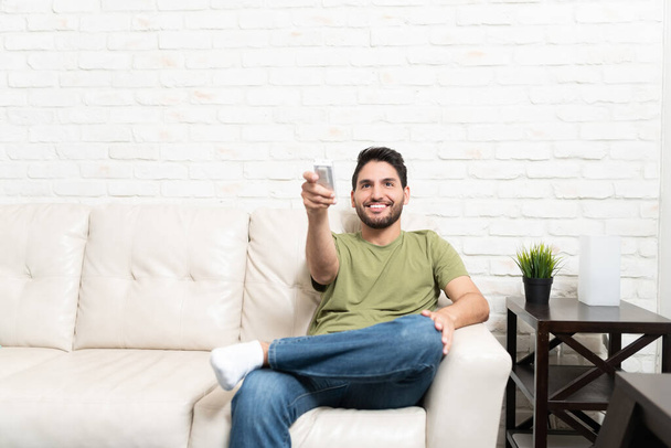 Усміхаючись гарним латиноамериканцем за допомогою кондиціонера дистанційного керування, сидячи на дивані біля цегляної стіни вдома. - Фото, зображення
