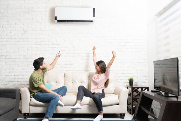 Boyfriend turning on air conditioner while cheerful girlfriend sitting with arms raised on sofa at home - Photo, Image