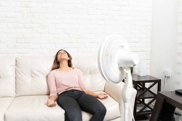 Латиноамериканская красивая молодая женщина охлаждается перед вентилятором, расслабляясь дома на диване
 - Фото, изображение