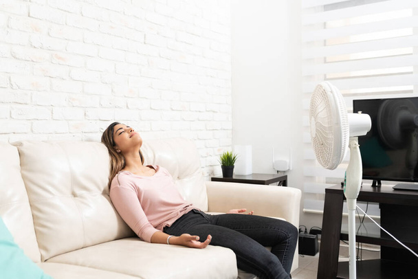 Bastante joven hispana enfriándose con ventilador mientras se relaja en el sofá en la sala de estar en casa
 - Foto, imagen