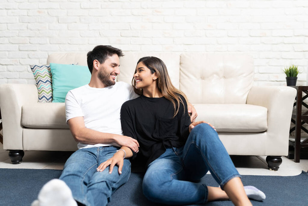 Feliz novio y novia hispanos en casuales relajándose contra el sofá en la sala de estar
 - Foto, Imagen