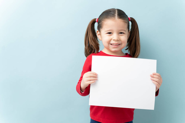 Portrait of a beautiful little girl with ponytails holding a white sign and smiling in a studio - Foto, Imagen