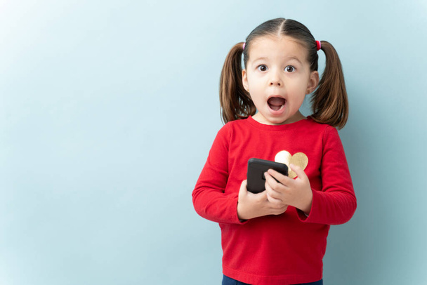 Portrait of a beautiful girl with ponytails holding a smartphone and looking shocked in a studio - Photo, Image