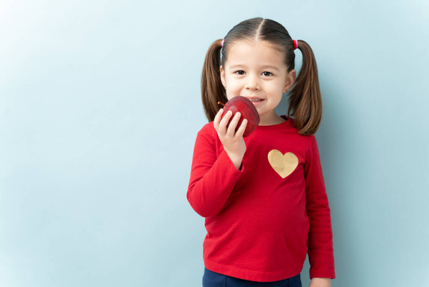 Маленька кавказька дівчинка з хвостами, що їдять яблуко і посміхаються в студії - Фото, зображення