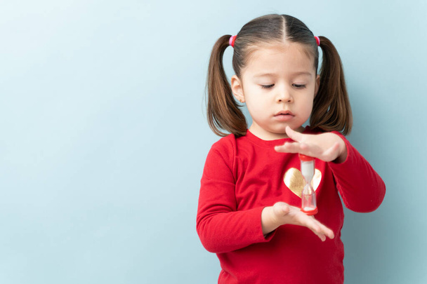 Serious looking little girl looking at a sand timer while practicing self-control during timeout in a studio with copy space - Φωτογραφία, εικόνα