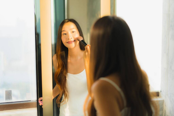 Retrato bela jovem mulher asiática compõem com pó cosmético e batom em seu rosto facial no interior do banheiro
 - Foto, Imagem