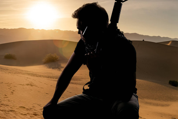 Silhueta de um soldado masculino descansando à sombra em um deserto e segurando uma espingarda. Representa a indústria militar privada, milícia ou forças especiais. Parece cansado e com saudades de casa.
 - Foto, Imagem