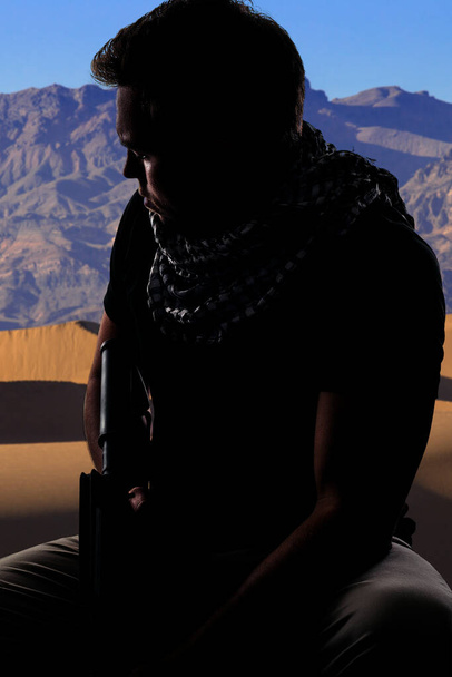 Silhouette of a male soldier resting in the shade on a desert and holding a rifle.  Depicts the private military industry, militia, or special forces.  He looks tired and homesick - Photo, Image
