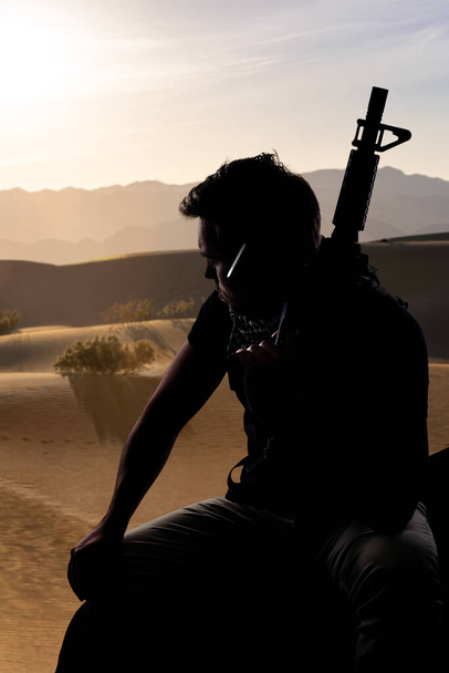 Silhouet van een mannelijke soldaat die rust in de schaduw van een woestijn en een geweer vasthoudt. Vertegenwoordigt de particuliere militaire industrie, militie of speciale troepen. Hij ziet er moe en heimwee uit. - Foto, afbeelding