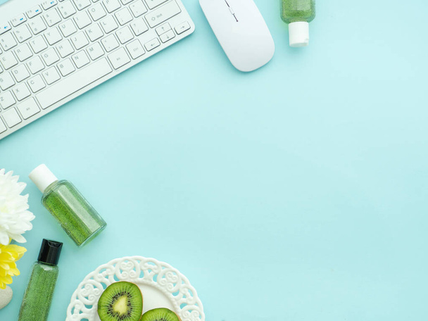 Freelancer woman blogger workplace, working space. Keyboard, mouse laptop, cosmetics shower gel bottles; kiwi fruits, peony flowers isolated on light blue table background with copyspace on the bottom - Foto, imagen