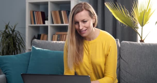 Cheerful woman sitting on couch and working on laptop - Záběry, video