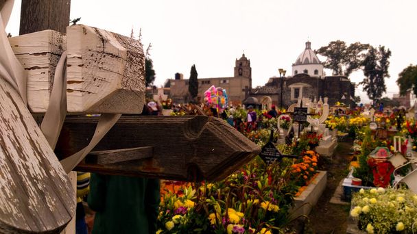 Primer plano de una cruz en un panten y al fondo las tumbas recin adornadas con flores muy coloridas y llamativas en un dia nublado y triste. - Foto, immagini