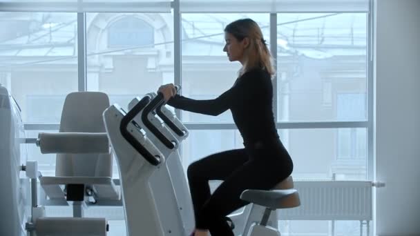 Uma jovem mulher senta-se em bicicleta de exercício no ginásio no fundo de grandes espelhos
 - Filmagem, Vídeo