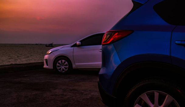 Großaufnahme Luxus-SUV und weißes Auto geparkt auf Betonparkplatz neben dem Strand am Abend mit schönen lila Sonnenuntergang Himmel. Roadtrip im Sommerurlaub am tropischen Meeresstrand.  - Foto, Bild