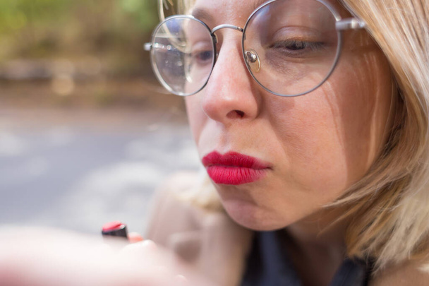 Kobieta maluje usta czerwoną szminką. Dziewczyna robi makijaż na ulicy. Portret blondynki w okularach. - Zdjęcie, obraz