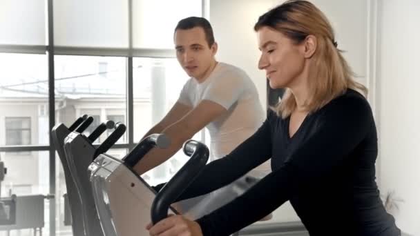 Hombre y mujer deportistas entrenando sus piernas en bicicleta estática y hablando entre sí
 - Imágenes, Vídeo