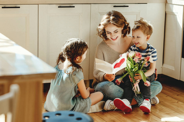 Улыбающаяся молодая женщина читает поздравительную открытку, сидя на полу с веселыми маленькими братьями и сестрами с букетом цветов во время мамы-мамы
 - Фото, изображение