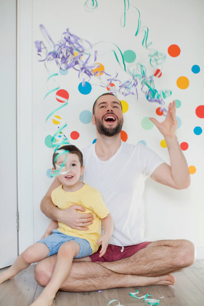 Mały chłopiec z ojcem w urodzinowych kapeluszach bawi się konfetti na tle ściany z kolorowymi kółkami - Zdjęcie, obraz