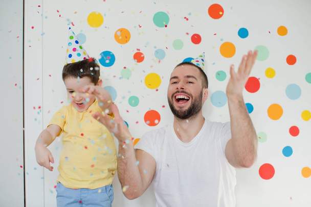 Kleinkind mit seinem Vater in den Geburtstagsmützen spielt mit Konfetti auf einer Wand mit bunten Kreisen - Foto, Bild