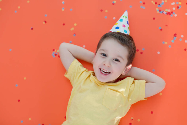 Enfant caucasien garçon dans le chapeau d'anniversaire sur un fond de papier rouge avec des confettis colorés
 - Photo, image