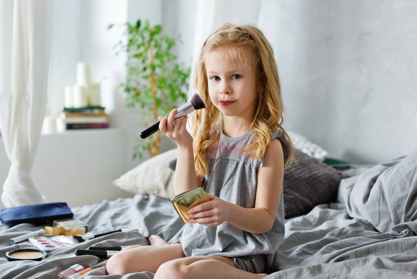 Happy child. Little cute girl doing makeup on the bed in the bedroom . The girl is laughing with makeup brushes in her hands - 写真・画像