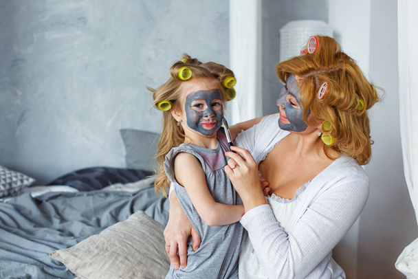 Glückliche Mutter und Tochter mit schwarzen Gesichtsmasken im Schlafzimmer auf dem Bett. Das Konzept der Heimunterhaltung. - Foto, Bild