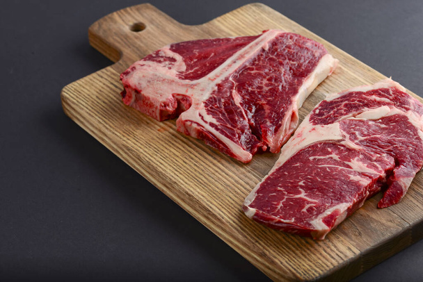 Fresh raw beef T-bone steak on wooden cutting board, top view. Porterhouse steak served over black background. - Фото, изображение