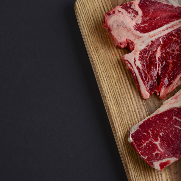 Fresh raw beef T-bone steak on wooden cutting board, top view. Porterhouse steak served over black background. - Photo, image