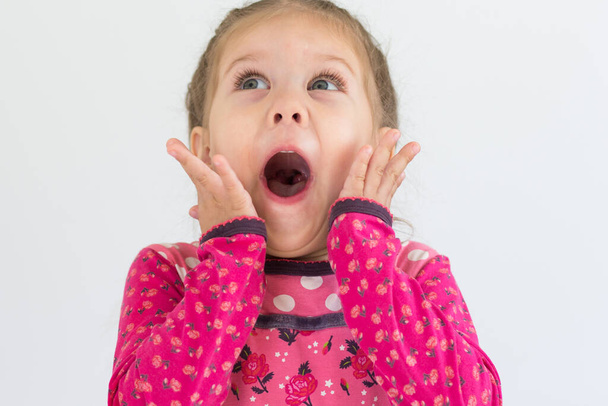 Portrait of caucasian child of three years old with surprised face holding hand close to open mouth looking up on white background - Zdjęcie, obraz