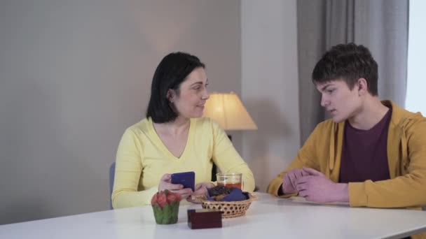 Young brunette Caucasian woman sitting at the table and using phone as teenage boy coming up and telling bad news. Son bringing shocking information to mother. Drama, problem, trouble. - Video, Çekim