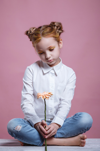 portret van een mooi klein meisje met bloemen. Studio shot op een roze achtergrond. Stijlvol en modieus klein meisje in een witte blouse en blauwe jeans - Foto, afbeelding