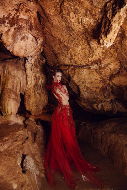 δυναμικό πλάνο μόδας. σέξι γυναίκα με κόκκινο φόρεμα, γοτθική νεράιδα γυρίσματα σε μια σκοτεινή σπηλιά. Κόκκινο σπήλαιο, Κριμαία - Φωτογραφία, εικόνα