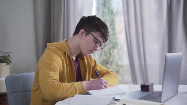 Camera moving from left to right around young Caucasian boy in eyeglasses writing and using laptop. Intelligent nerd student doing homework at the table. Education, intelligence, lifestyle. - Séquence, vidéo