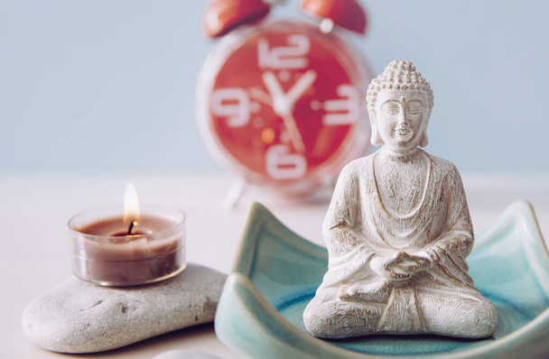 Időt találni az öngondoskodásra és az egyensúlyra a test lelke és elméje között. Egyensúly a munka és az otthon fogalma között. Lotus póz fehér szín ül meditáció Buddha figura, piros ébresztőóra a háttérben. - Fotó, kép