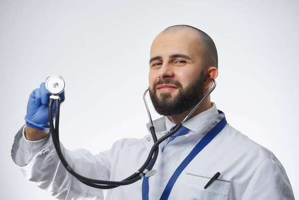Šťastný doktor se stetoskopem v ruce v modré lékařské rukavici. Lékař s plnovousem v bílém laboratorním plášti a modrou kravatou. - Fotografie, Obrázek
