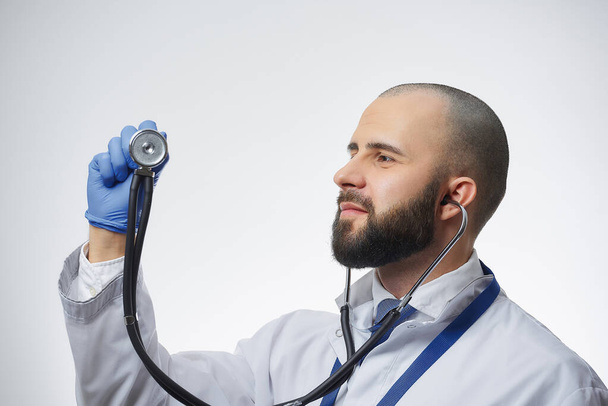 Szczęśliwy lekarz noszący stetoskop patrzący na jego głowę. Lekarz z brodą w białym fartuchu i niebieskim krawacie.. - Zdjęcie, obraz