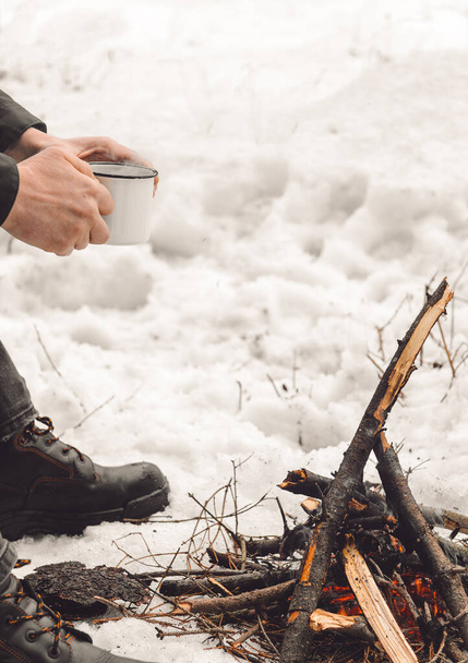 Male hands hold a mug of coffee near a burning campfire. Concept hike, walk, trip in winter - Photo, Image